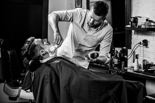 Client during beard shaving in barbershop. Young handsome barber and attractive bearded man. Black and white or colorless photo. Hairstyle, salon, hairdresser, barber shop, lifestyle concept