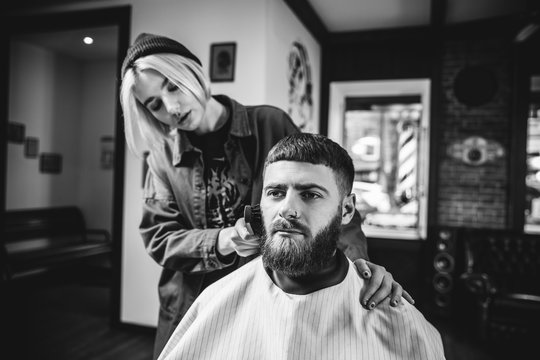 Young woman making haircut for bearded man at barbershop. Female barber at salon. Gender equality. Woman in the male profession. Black and white or colorless photo. Hairstyle, salon, hairdresser