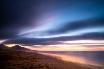 Obraz premium Beautiful landscape in long exposure at the beach during coloured cloudy sunset with mountains in background - wild and scenic vacation concept - cute outdoor nature with colors and ocean horizon