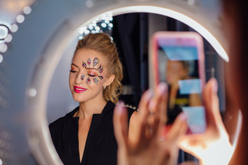 Beautiful young woman with flower face art. Spring makeup. Creative woman holiday make-up with sparles and decorative crystals. Woman taking portrait picture with mobile phone.