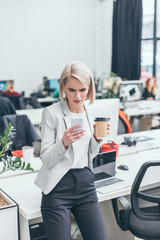 pretty serious businesswoman using smartphone and holding disposable cup in office