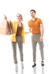 Obraz premium pretty smiling woman holding paper bags and pointing with finger while standing near cheerful man isolated on white