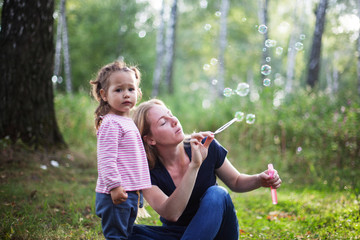 Fototapeta premium Mother and little daughter blowing soap bubbles in park. Happy and carefree childhood. Good family relations.