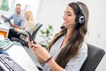 Friendly female customer support operator with headset using phone in office