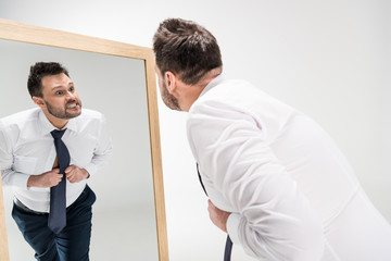 angry overweight man in formal wear looking at reflection in mirror on white