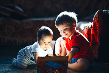 Children reading a book sitting together on floor at home. brother and babysister smiling having fun with book together. Boy and girl reading by the light of a sunset back light mode