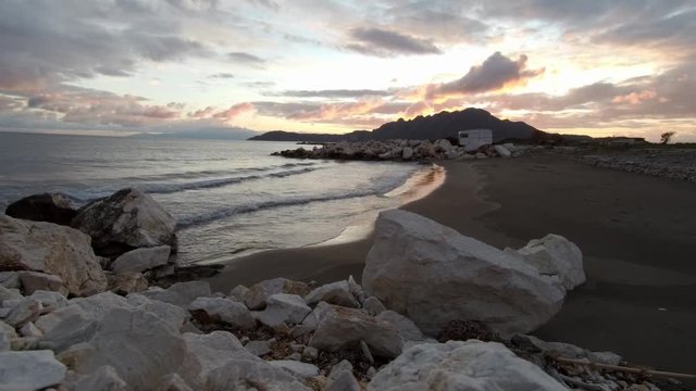 Wide shot of a sunset. Big rocks and a small beach with a camping trailer. Calm atmosphere waves slowly crashing on the shore