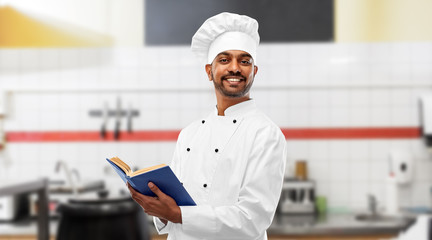 cooking, profession and people concept - happy male indian chef in toque with cookbook over restaurant kitchen background