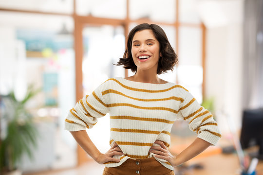 people concept - happy smiling young woman in striped pullover with hands on hips over office room background