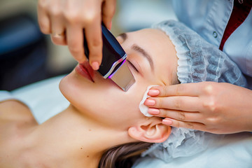 Obraz na płótnie Canvas Woman receiving ultrasonic facial exfoliation at cosmetology salon. Procedure clearing clogged pores, ultrasonic treatment for skin rejuvenation, beautician uses modern apparatus for refreshing