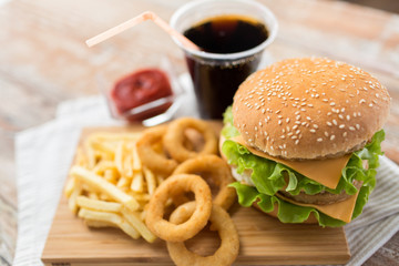 fast food and unhealthy eating concept - close up of double hamburger or cheeseburger, deep-fried squid rings, french fries, cola drink and ketchup on wooden board