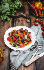 salad with roasted beef, tomatoes, lettuce and grilled pepper on white plate with fresh and grilled vegetables on brown wooden table, copy space