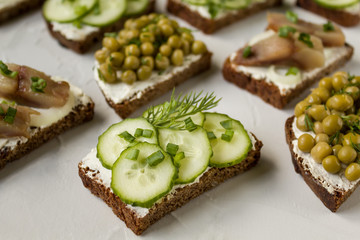 Hearty sandwiches of black bread with cucumbers, canned peas, salted fish on a gray background. Side view, close-up