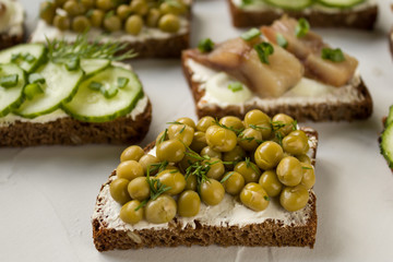 Hearty sandwiches of black bread with cucumbers, canned peas, salted fish on a gray background. Side view, close-up