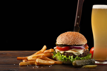 Homemade hamburger with french fries and glass of beer on wooden table. In the burger stuck a knife. Fastfood on dark background - obrazy, fototapety, plakaty