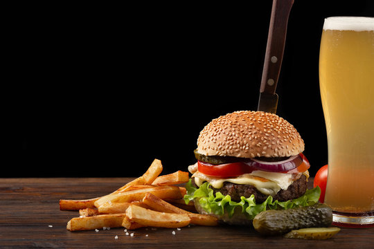 Homemade hamburger with french fries and glass of beer on wooden table. In the burger stuck a knife. Fastfood on dark background
