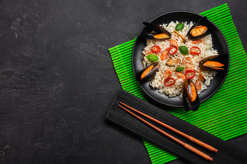 Fried rice with seafood mussels, shrimps and basil in a black plate with chopsticks on green bamboo mat and stone table