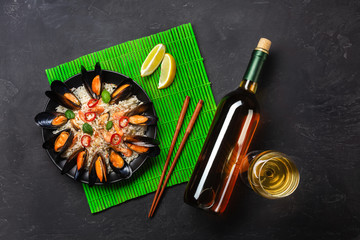 Fried rice with seafood mussels, shrimps, basil in a black plate with wine bottle, wineglass, lemon, chopsticks on green bamboo mat and stone table