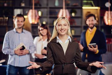 Young smiling blonde Caucasian businesswoman sredded elegant posing with her successful team in restaurant.