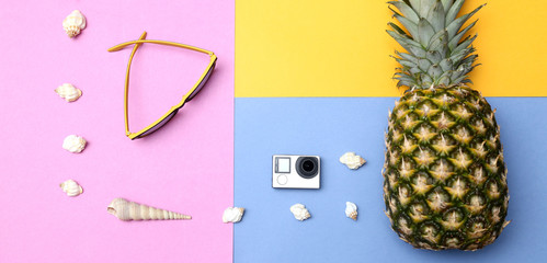 Pineapple, sunglasses, straw hat, action camera and seashells on a yellow blue pink background. Tropical summer vacation concept with pineapple.