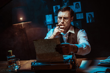 Detective in glasses smoking cigar while using typewriter in dark office