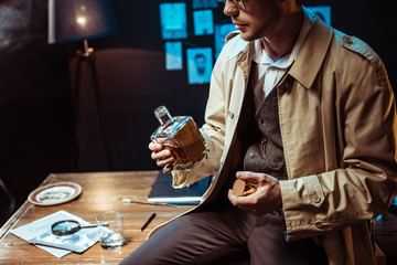 Cropped view of detective in trench coat sitting on table and holding bottle of cognac