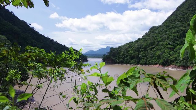 Time lapse from behind some plants, of the Amazon River in Bolivia, with a mountain in the background, on a sunny day with clouds passing by
