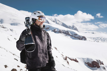 Portrait bearded male skier aged against background of snow-capped Caucasus mountains. An adult man wearing ski googles mask and helmet skis on his shoulder looks mountains. Ski resort concept