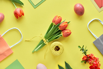 Easter flat lay on yellow paper. Bunch of tulips, gift boxes, decorative eggs and paper bags, geometric arrangement