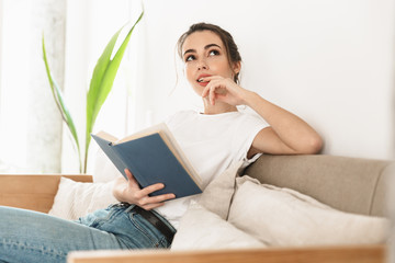 Beautiful young student girl sitting indoors reading book on sofa.