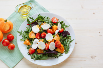 Fresh salad with arugula, cherry tomatoes, mozzarella cheese and olive oil on white wooden background and green napkin. Top view