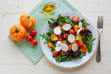 Fresh salad with arugula, cherry tomatoes, mozzarella cheese and olive oil on white wooden background and green napkin. Top view
