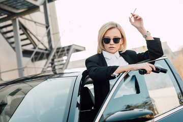 beautiful blonde smoker in sunglasses holding cigarette and gun near automobile