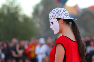 A side profile view of a young female Caucasian dancer wearing a white mask at an outside event.