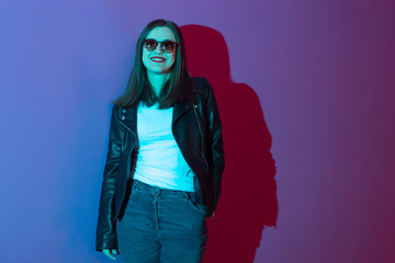 Fashioned girl in leather jacket in dark glasses in pink anf blue  neon lights background. 