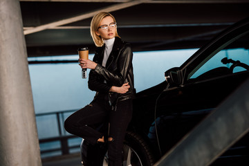 Fototapeta na wymiar beautiful blonde girl in glasses holding paper cup and key in parking near car