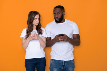 Mistrustful interracial couple in white T-shirts holding smartphones in arms