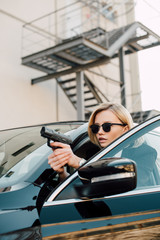 serious blonde woman in sunglasses holding gun near black car