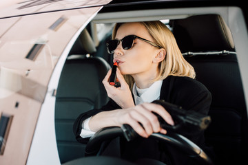 Fototapeta na wymiar attractive blonde woman applying lipstick while holding gun in car