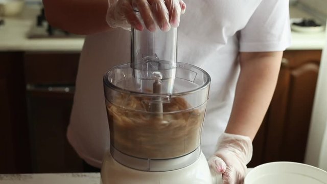 Young housewife chops peeled walnuts in a kitchen combine in the kitchen. Women's hands in white transparent culinary gloves. Vegan food. Close-up.