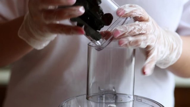 A young housewife pours prunes from a glass cup into a food processor in the kitchen. Women's hands in white transparent culinary gloves. Vegan food. Close-up.