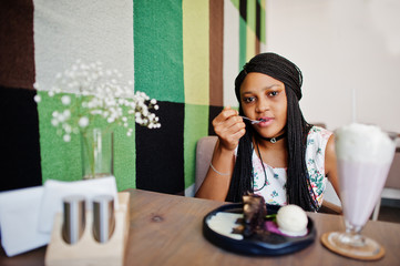 Сheerful african american young woman in summer dress and choker necklace at cafe eating chocolate cake with ice cream.