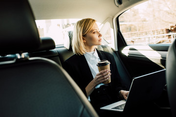 Fototapeta na wymiar beautiful blonde woman holding paper cup near laptop in car