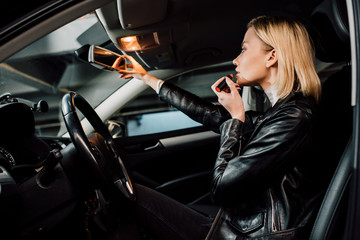 Fototapeta na wymiar low angle view of attractive blonde girl applying lipstick and touching mirror in car