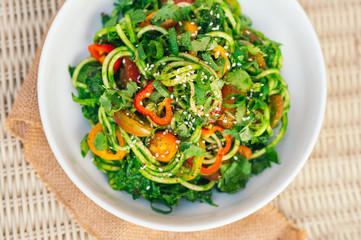 Zucchini spaghetti salad with yellow tomatoes, red pepper, onion, seasame seeds, cilantro in white bowl. Fresh salad on table. Vegetable dish. Horizontal top view shot