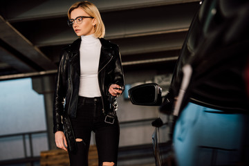 attractive girl in glasses holding key in parking near black automobile