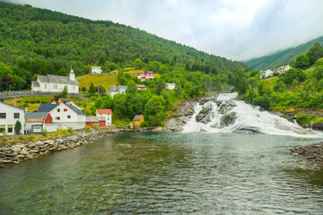 Fototapeta na wymiar Houses on mountainside by lake near the waterfall. Mountain river flows into lake from top of mountain. Hellesyltfossen is one of many Norwegian waterfalls in area Geirangerfjord in village Hellesylt.