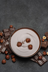 glass bowl of chocolate cream or melted chocolate, pieces of chocolate and hazelnuts on dark concrete background