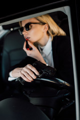 Plakat selective focus of gun in hand of attractive blonde woman applying lipstick in car