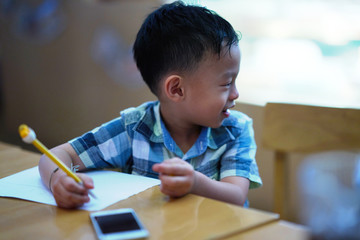 Boy is writing paper with pencil and parents smart phone on table with fun
